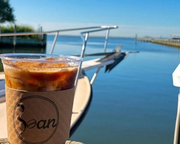 An iced coffee in a Bean sleeve on the dock, with boats and calm water behind it